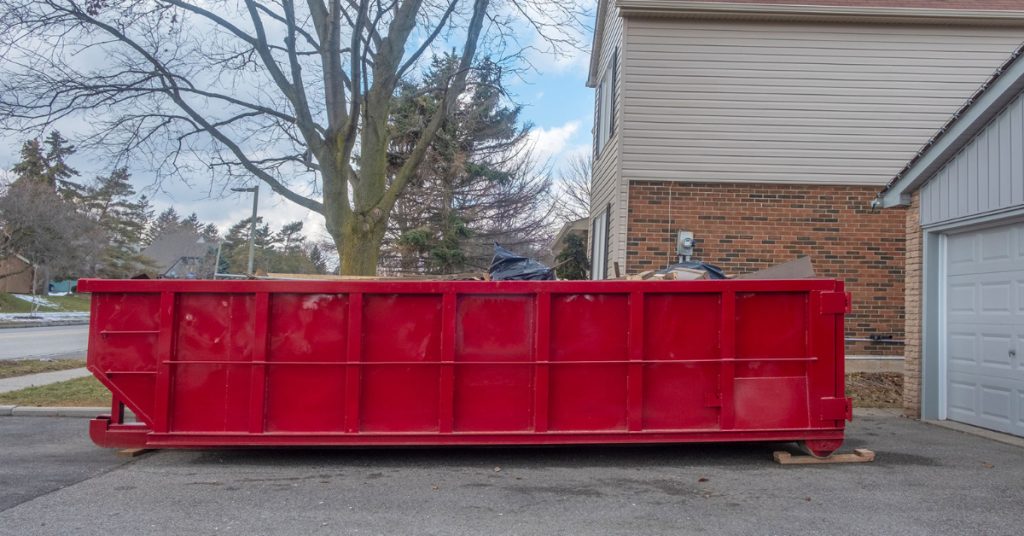 A red dumpster full of debris from KEI dumpster rental services sits in the driveway of a home. The homeowner placed wooden skids under the dumpster to protect the driveway from damage. 