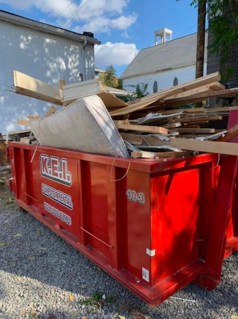 A red KEI roll-off dumpster that's overloaded with construction debris and other materials. 