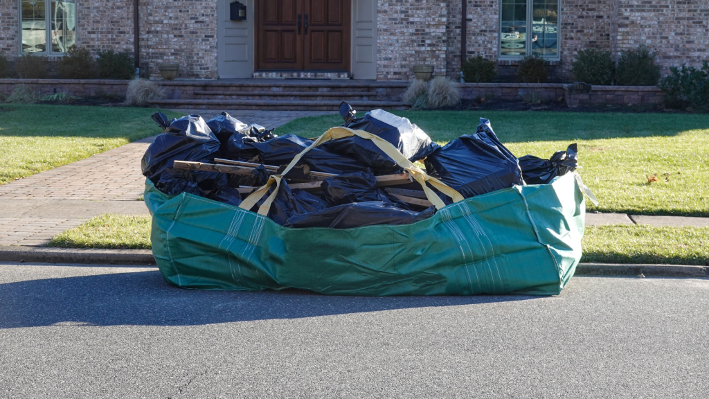 A Bagster disposal system sits at the curb loaded with black garbage bags full of debris. 