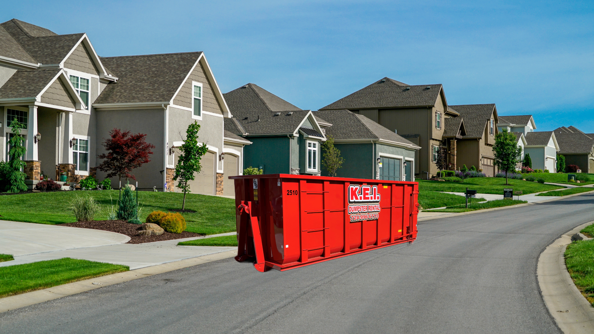 A red KEI roll-off dumpster sits curbside in a residential neighborhood. The dumpster is in the street, sandwiched between two driveways.