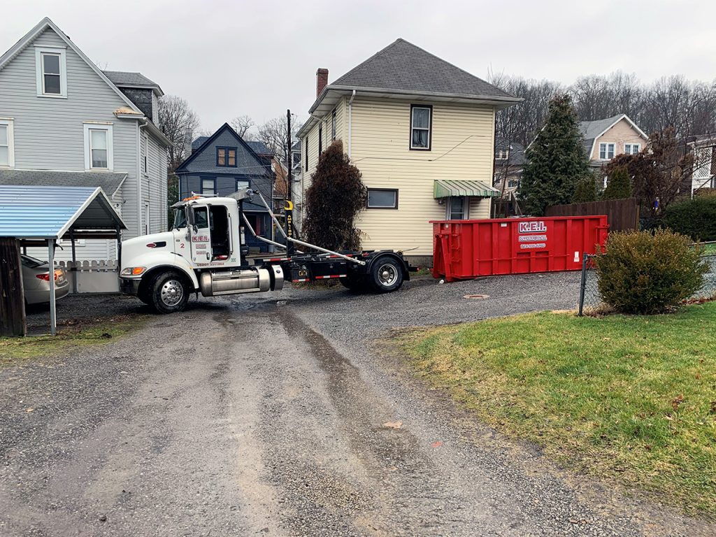 A white KEI truck delivers a large red KEI roll off dumpster to a residential neighborhood. 
