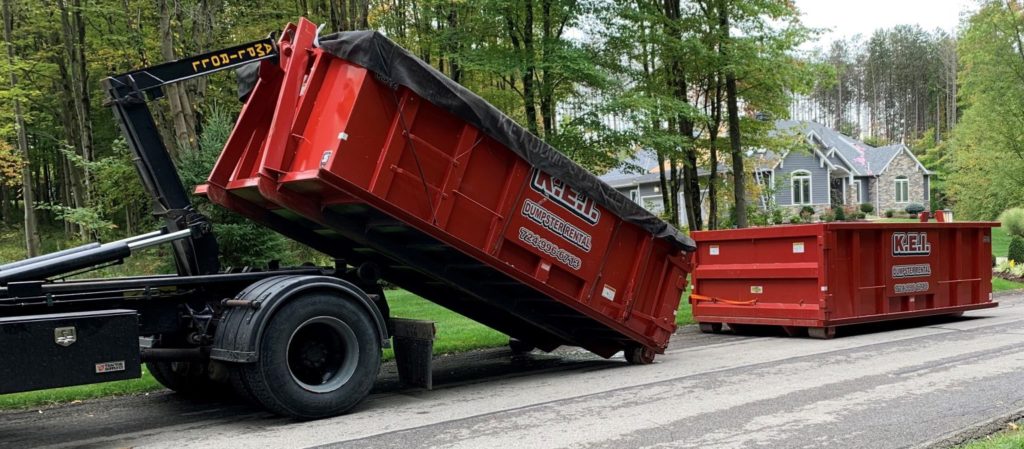 A KEI truck delivers two red roll-off dumpsters to a home in a neighborhood. 